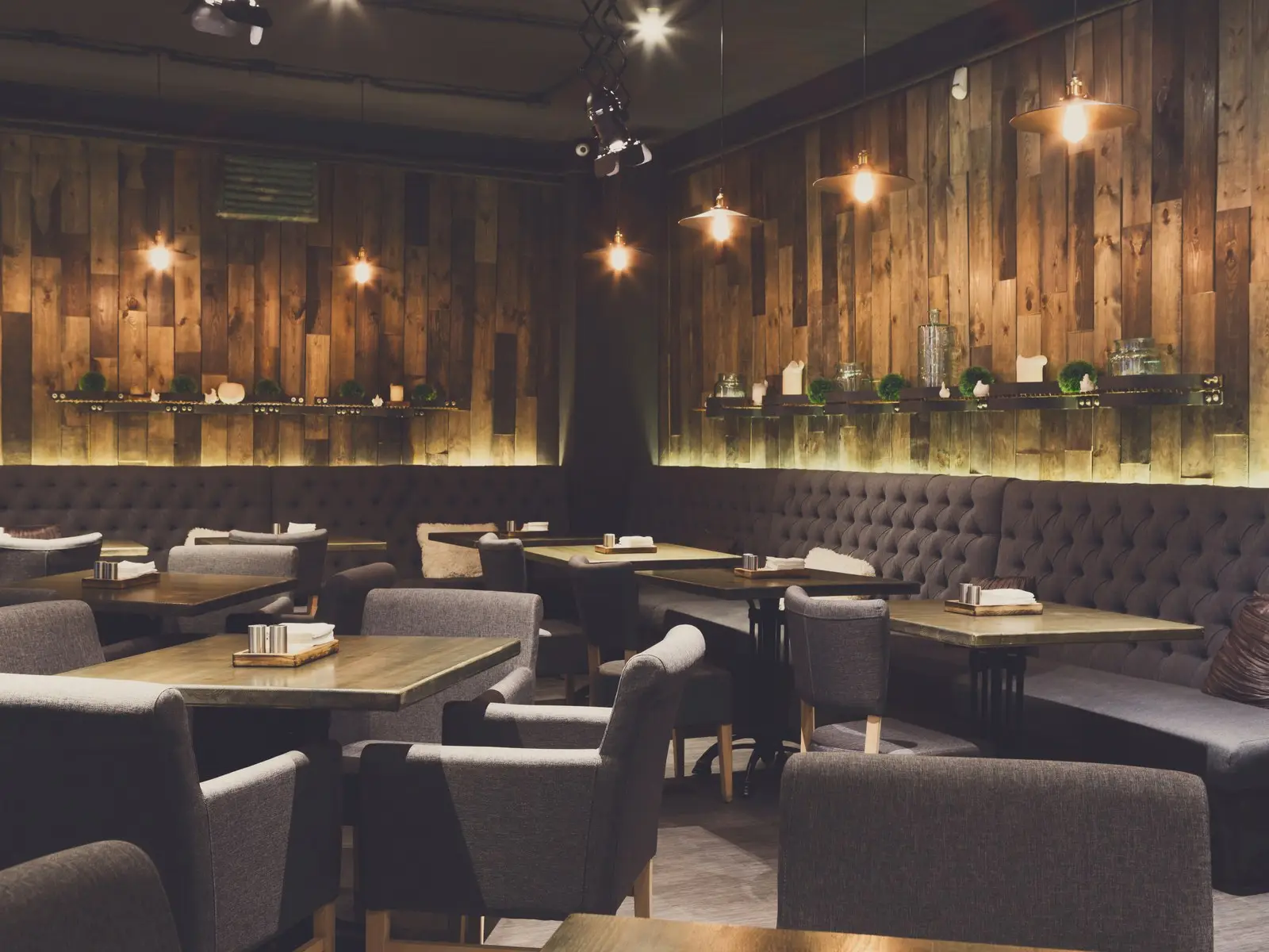 Salle de restaurant chaleureuse avec mur en bois, banquettes capitonnées gris foncé et suspensions lumineuses.