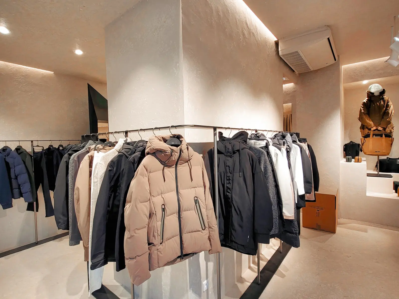 Portants métalliques avec vestes autour d'un poteau central dans une boutique aux murs enduits beige et éclairage chaud.