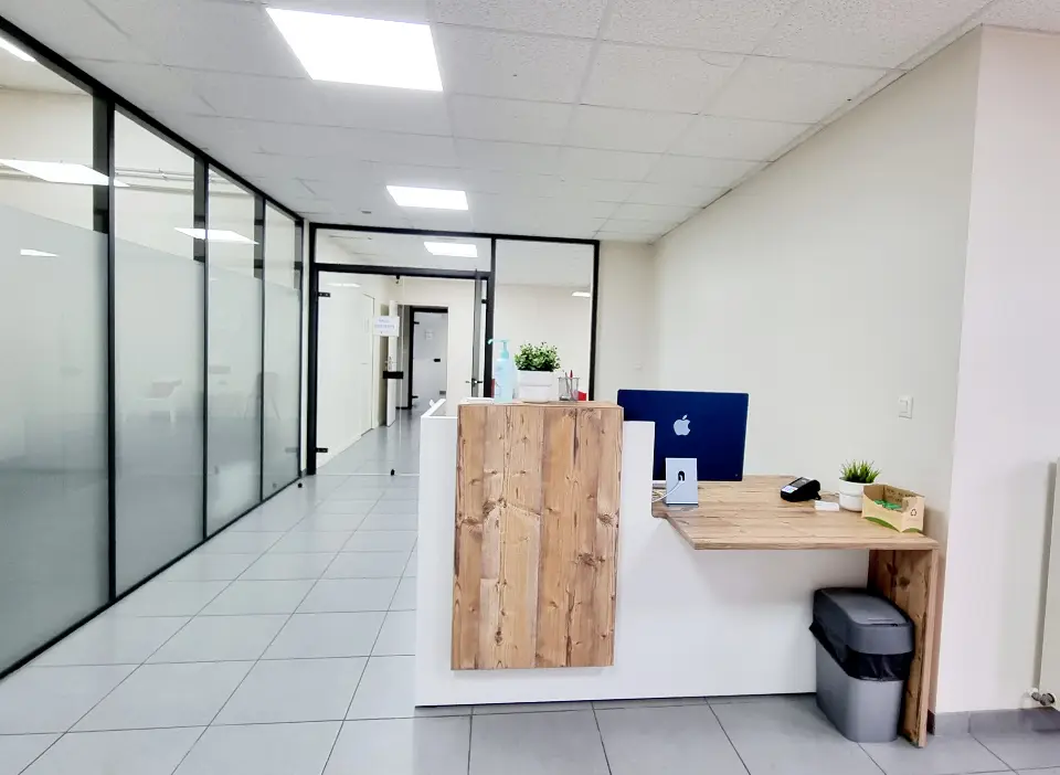 Un comptoir de réception en bois clair et blanc au milieu d'un couloir de locaux professionnels lumineux, avec des boîtes de rangement.