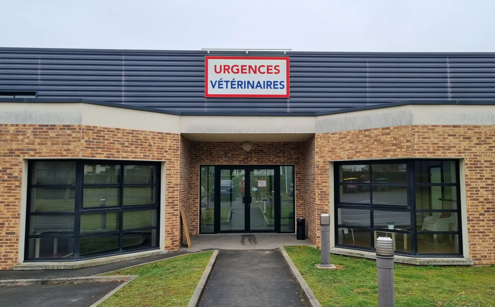 Façade avant d'une clinique vétérinaire avec briques oranges, bardage gris et enseigne "Urgences Vétérinaires" rouge et bleue.