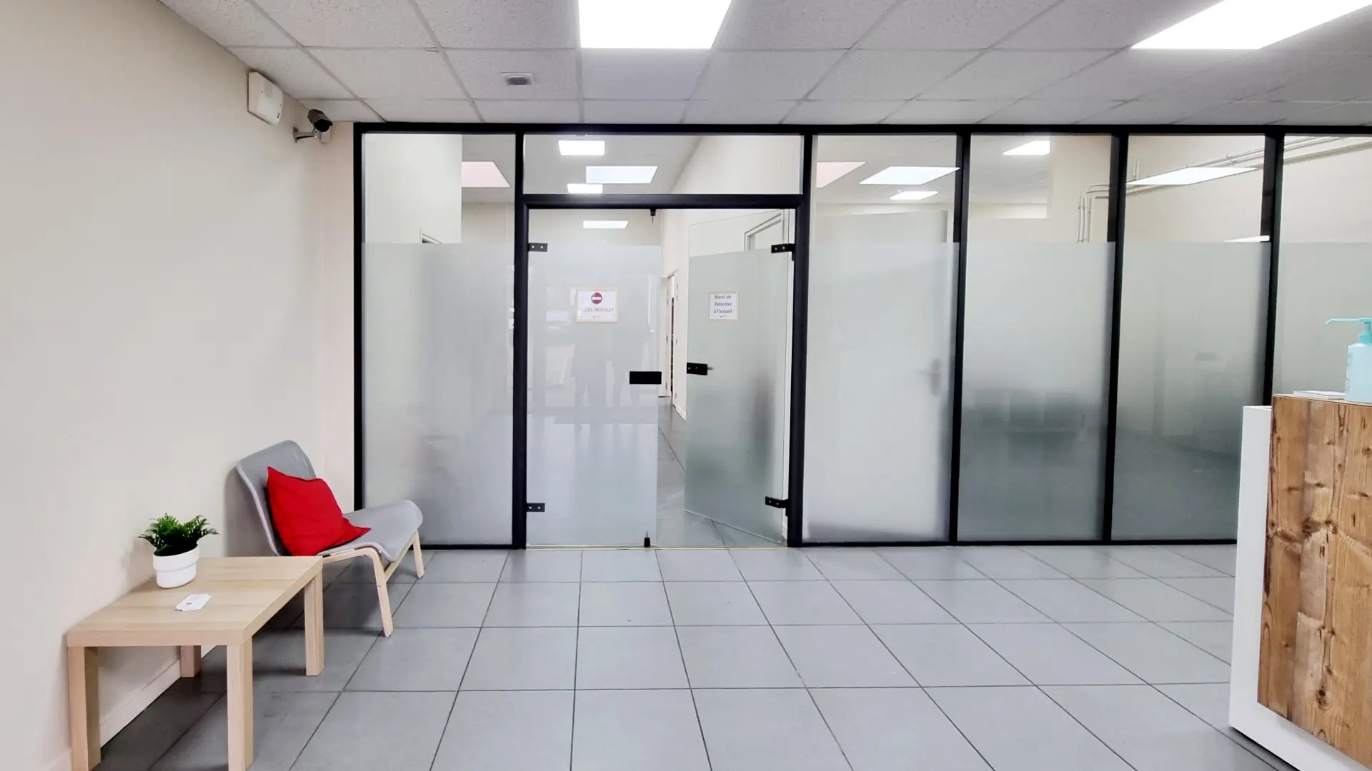 Vue d'une salle d'attente lumineuse avec un mur crème, des carreaux gris, et un mur vitré. Une petite table en bois clair, un fauteuil gris avec un coussin rouge se trouvent sur la gauche. L'entrée vitrée est dotée d'une signalétique contrastée.