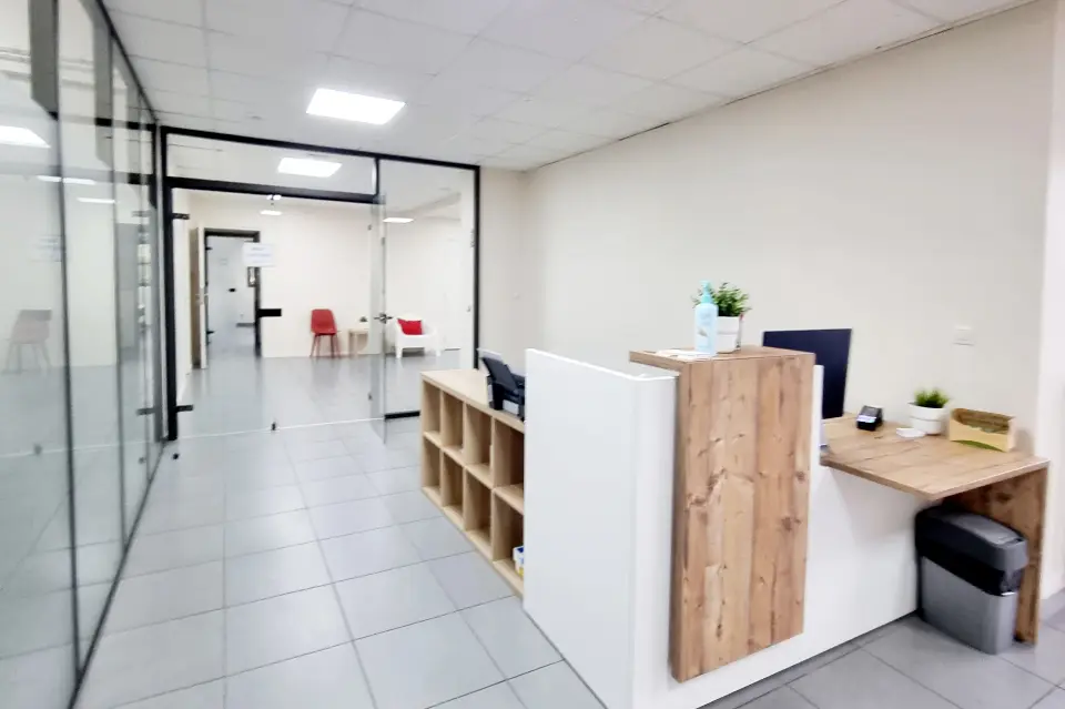 Un comptoir d'accueil en bois clair et blanc, avec un plafond carrelé, et des bureaux vitrés et des zones d'attente. Lumière naturelle et artificielle.