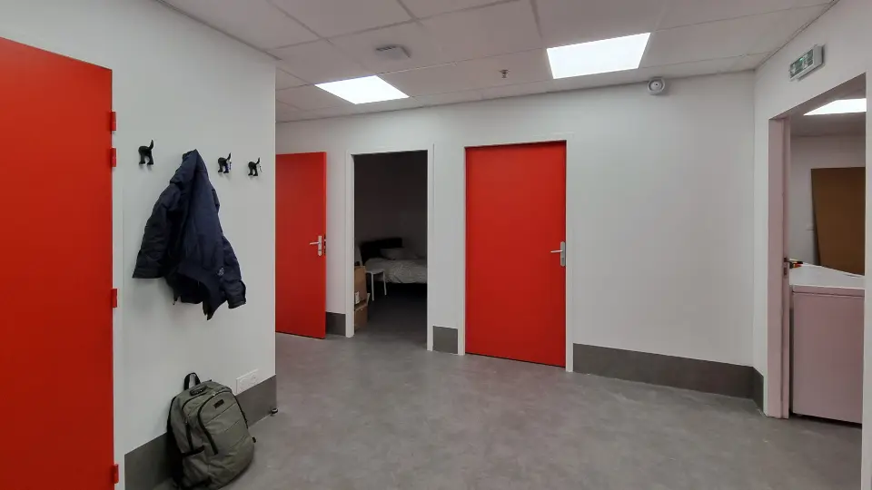Couloir moderne, lumineux, aux murs blancs et sol gris, avec des portes rouge vif et un manteau sur patères murales.