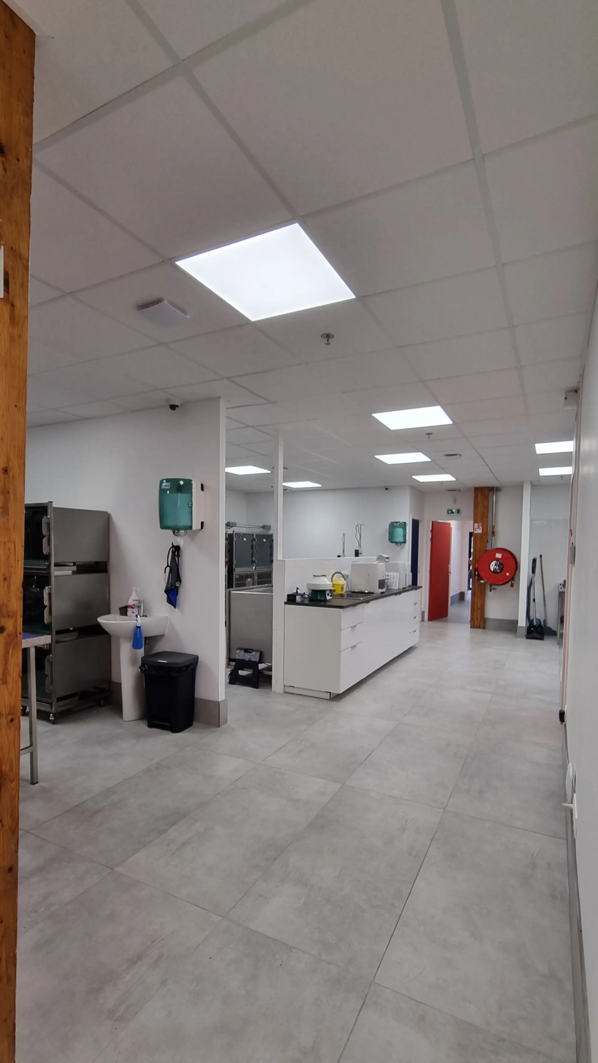 Vue large d'un couloir de clinique vétérinaire moderne avec des murs blancs, un sol gris clair et des luminaires encastrés. Un évier est visible à gauche.