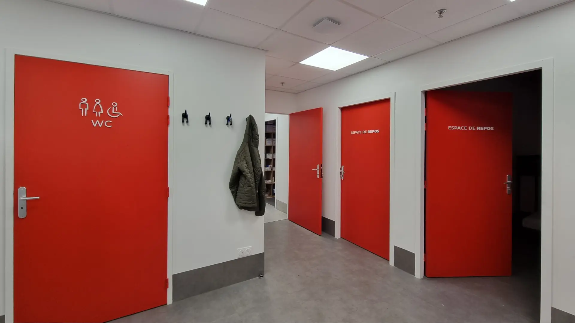 Couloir lumineux avec des portes rouges donnant sur les toilettes et deux "Espace de repos". Murs blancs et sol gris.