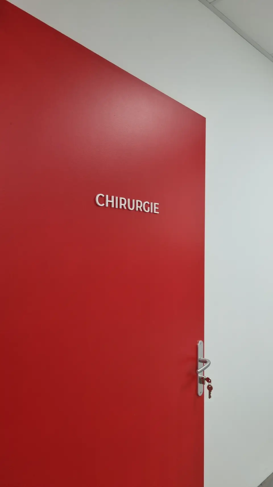 Vue en plongée sur une porte rouge vif marquée "CHIRURGIE", avec un espace blanc juste au-dessus.