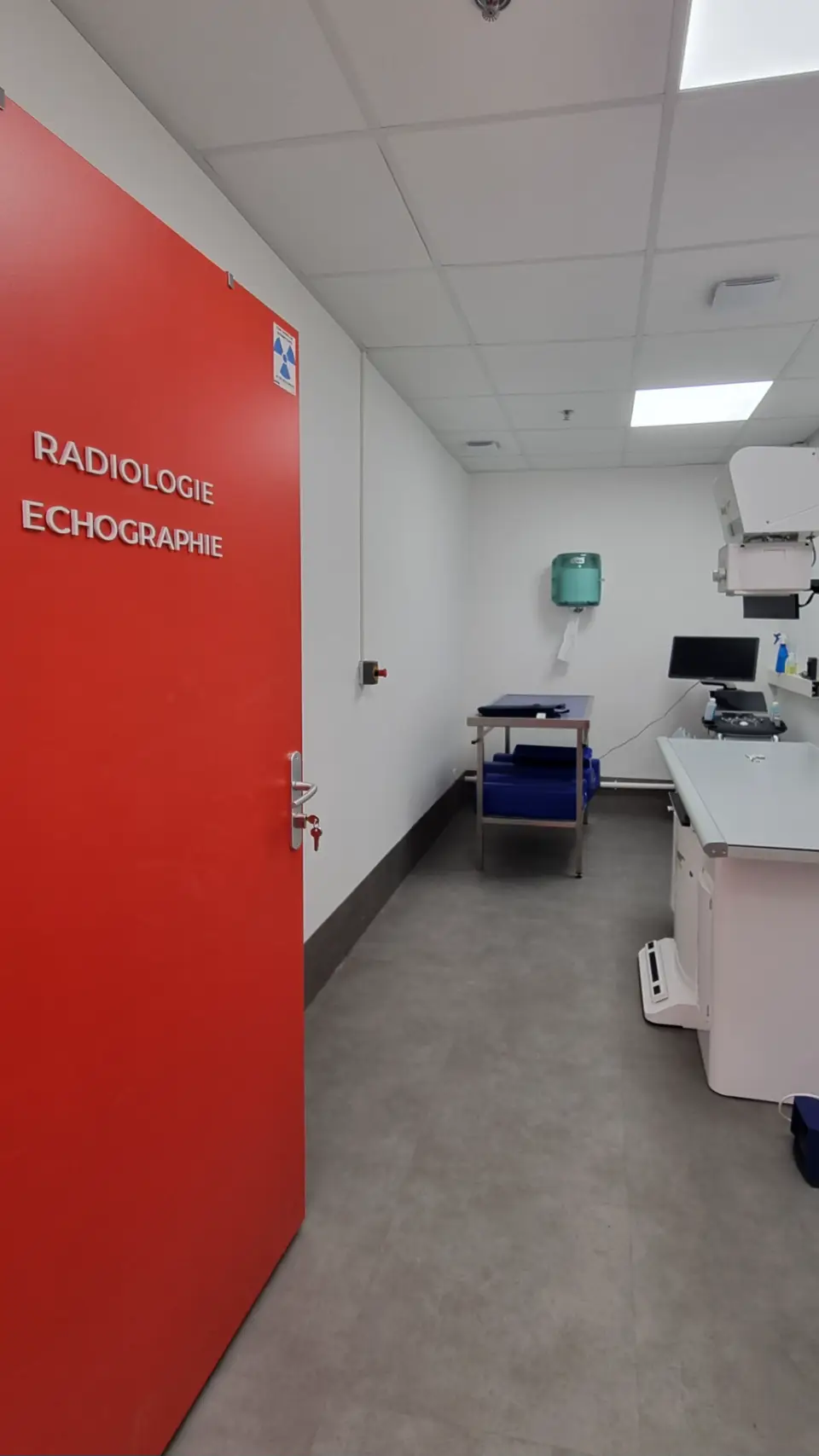 Un couloir lumineux dans une clinique vétérinaire, avec une porte rouge vif menant à la salle de radiologie et d'échographie, des surfaces blanches, et des équipements médicaux modernes disposés le long d'un mur.