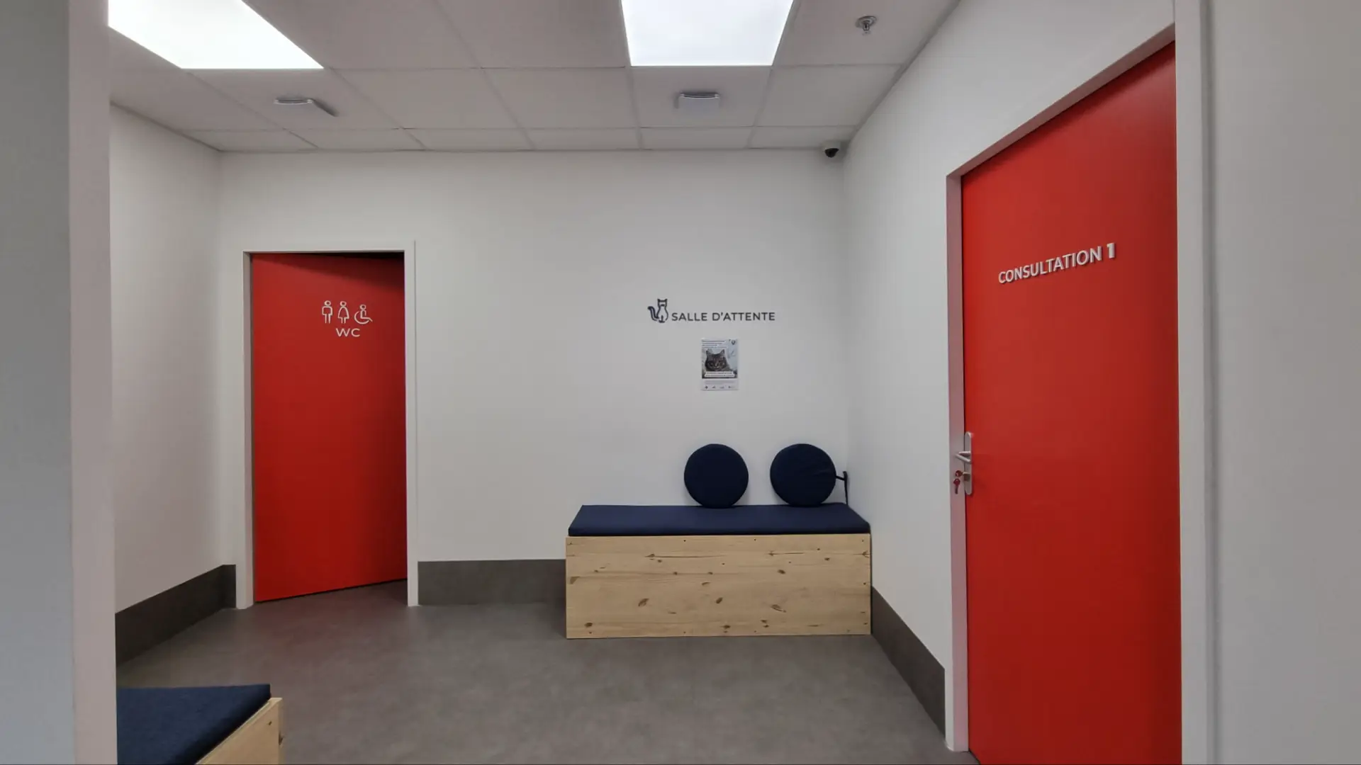 Couloir lumineux avec trois portes rouges vives dont une avec "WC" et une autre "CONSULTATION 1", et une banquette en bois clair avec coussin bleu foncé.
