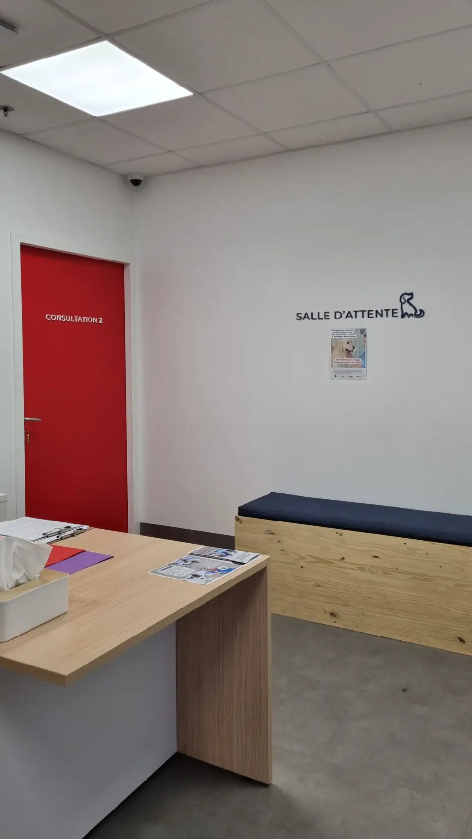 Vue d'une salle d'attente lumineuse avec un comptoir en bois clair, une porte rouge et un banc en bois.