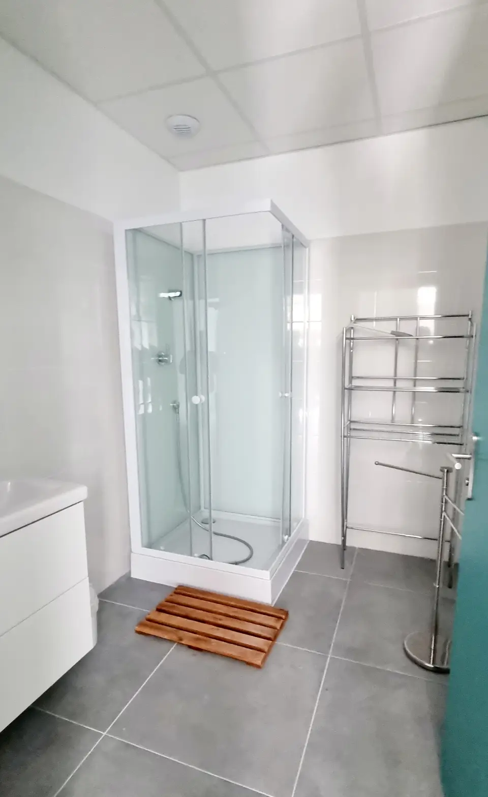 Salle de bain moderne et lumineuse avec douche vitrée, meuble-vasque blanc, sol carrelé gris et porte bleue, lumière zénithale.