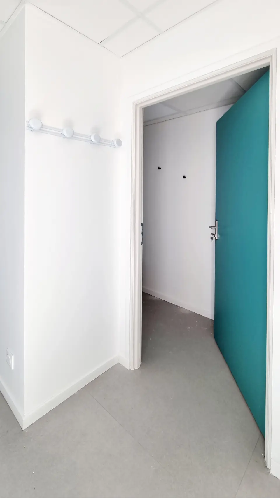 Un couloir blanc et épuré mène à une porte turquoise ouverte, révélant une autre pièce claire.