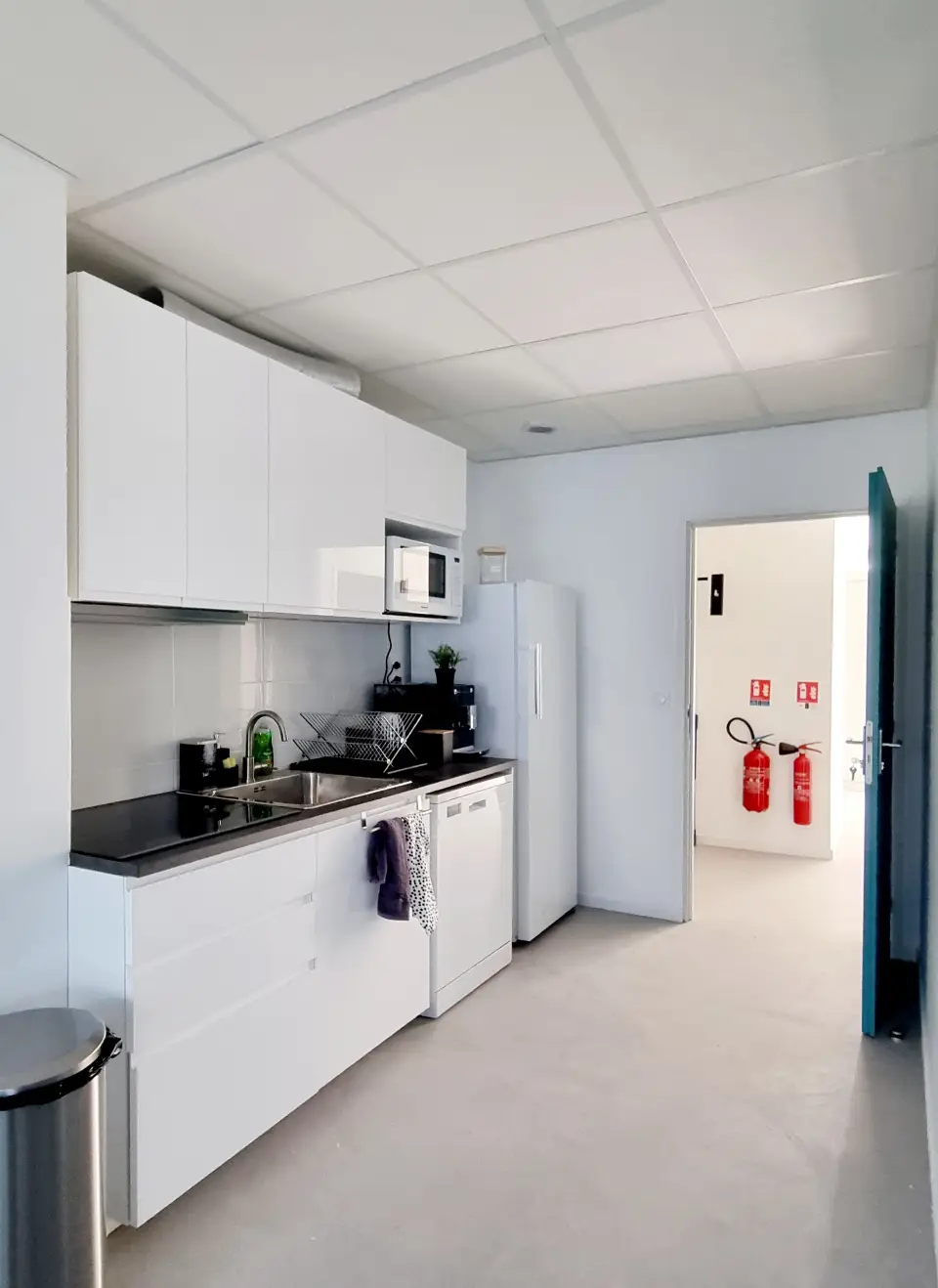 Cuisine moderne avec des armoires blanches brillantes, un comptoir noir, un évier en inox et un réfrigérateur. Sol en béton ciré gris.