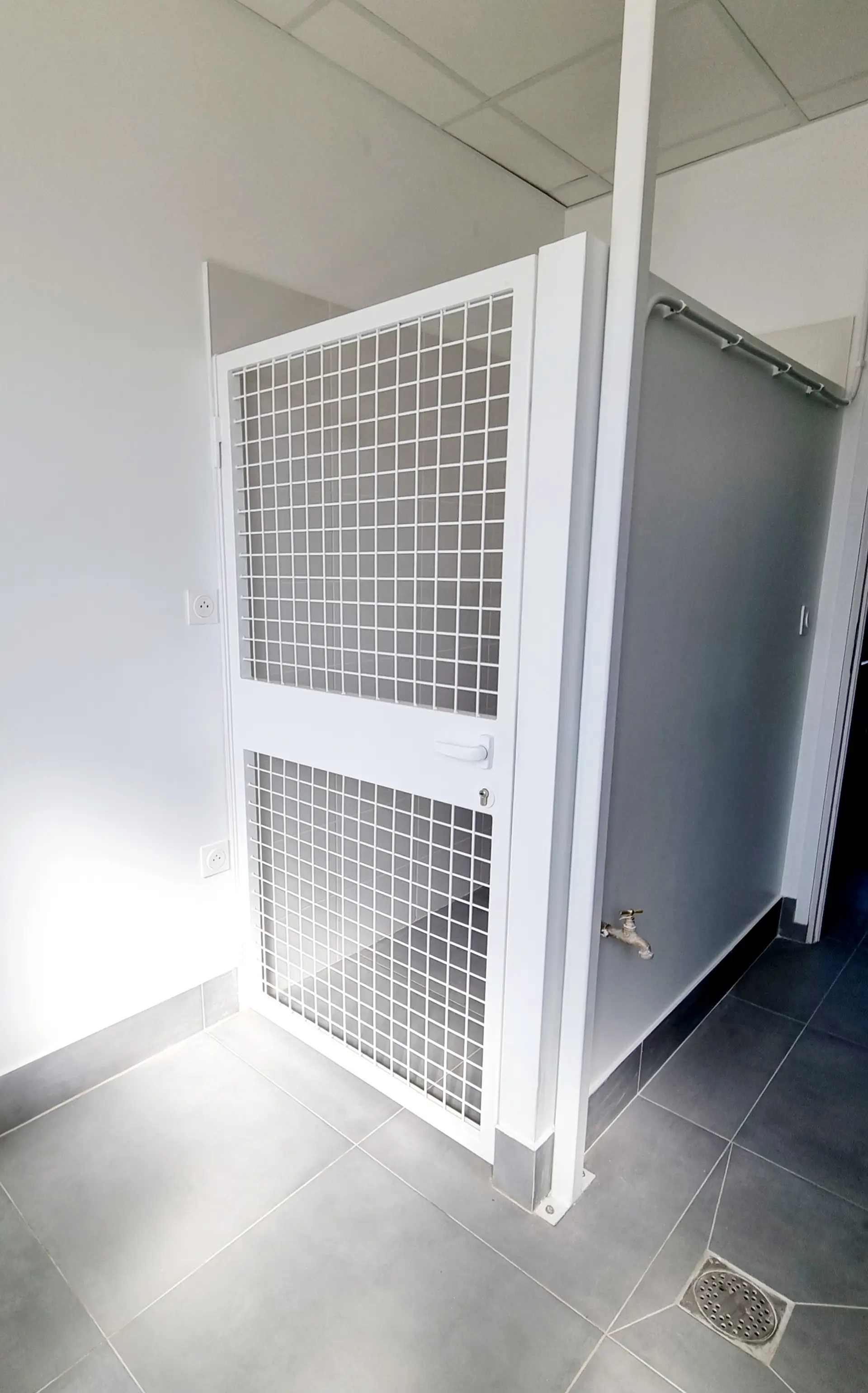 Un box d'hébergement pour animaux avec une porte grillagée blanche, un carrelage gris et un mur gris.
