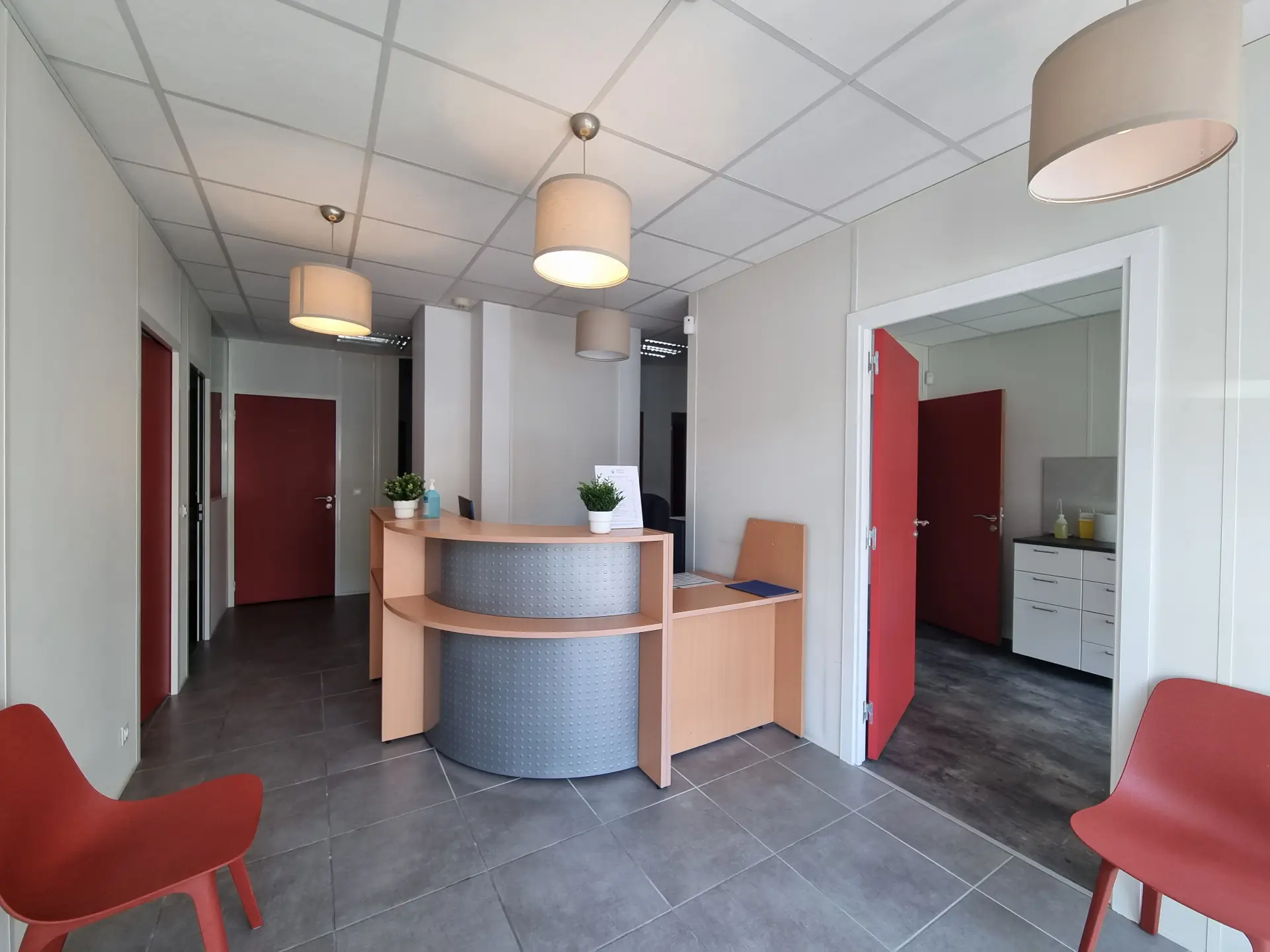 Vue en perspective d'un couloir de salle d'attente baigné de lumière, mobilier en bois clair, chaises rouges, portes rouges, carrelage gris.