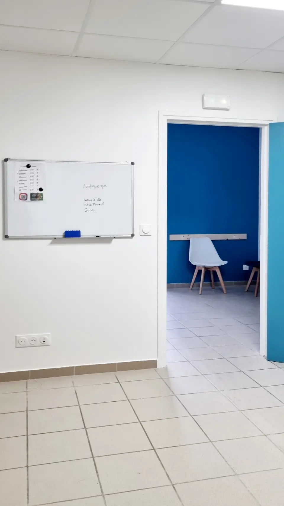 Un couloir blanc et carrelé avec tableau blanc et prise électrique, menant à une pièce aux murs bleus et chaise moderne.