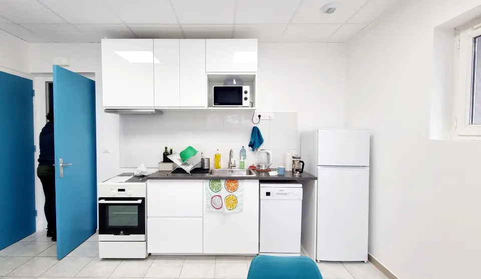 Cuisine moderne avec des meubles hauts et bas blancs, un plan de travail sombre, et des appareils électroménagers blancs. Une personne est derrière une porte bleue ouverte à gauche.