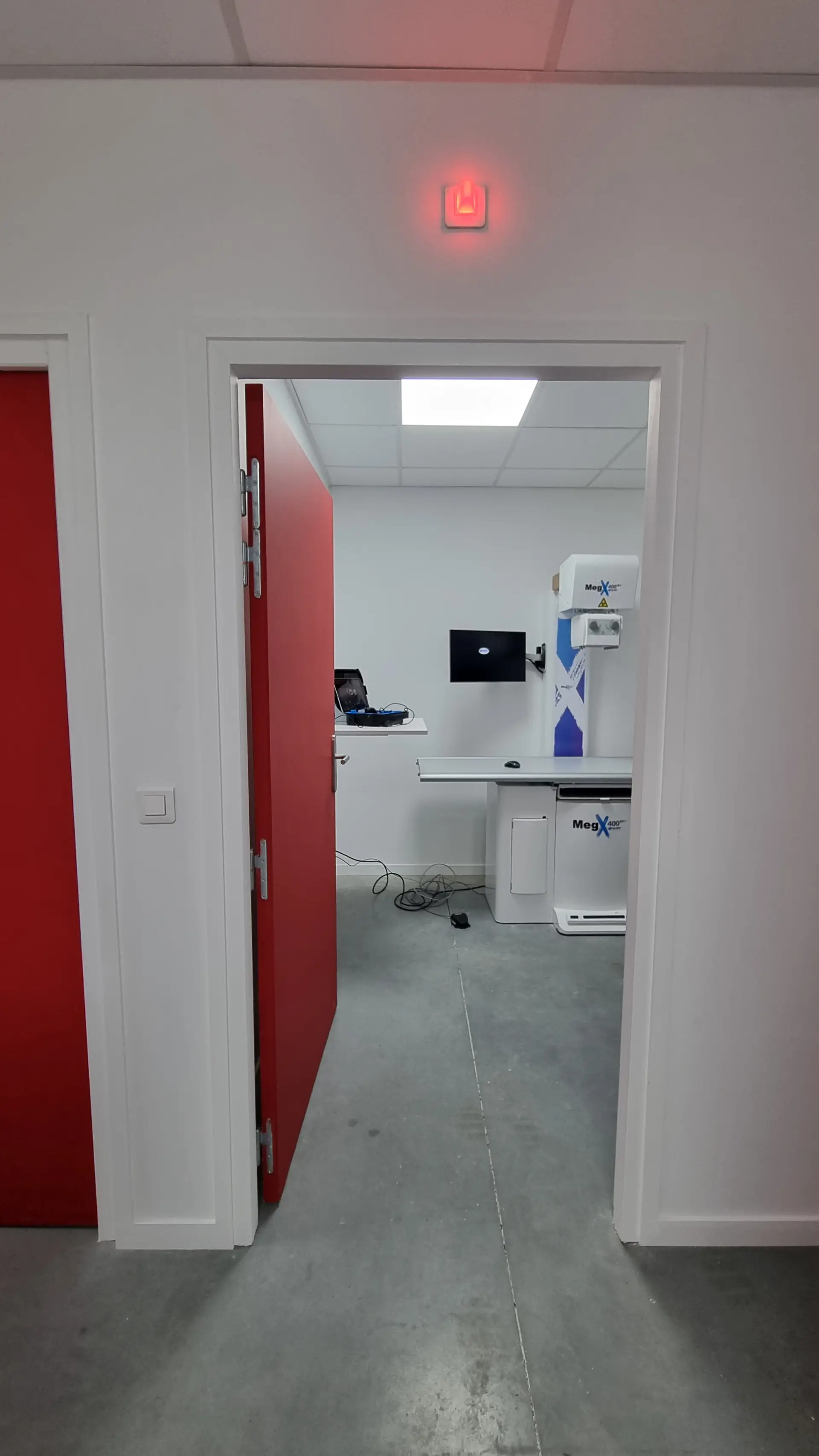 Vue à travers une porte entrebâillée rouge sur une salle d'examen vétérinaire moderne avec équipement de radiographie et écran.