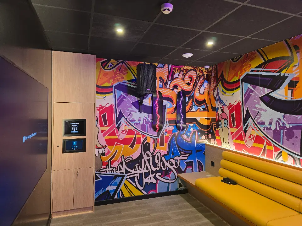 Salle de karaoké moderne avec mur graff, écran géant, meuble média bois clair et banquette jaune vif éclairée.