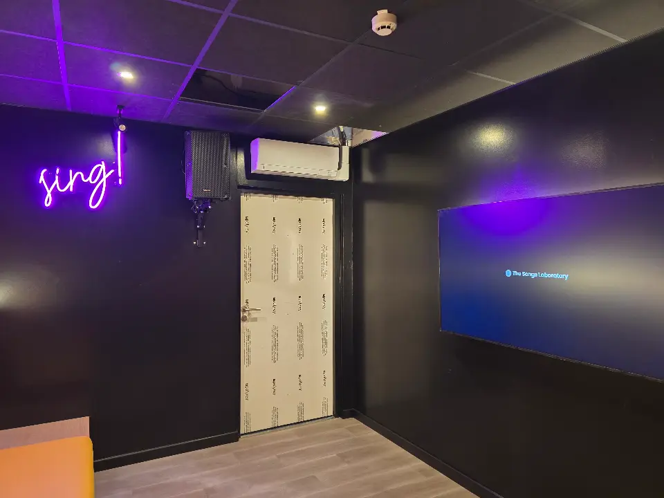 Salle de karaoké moderne avec murs noirs, enseigne néon violette "sing!", écran TV, porte claire, sol stratifié.