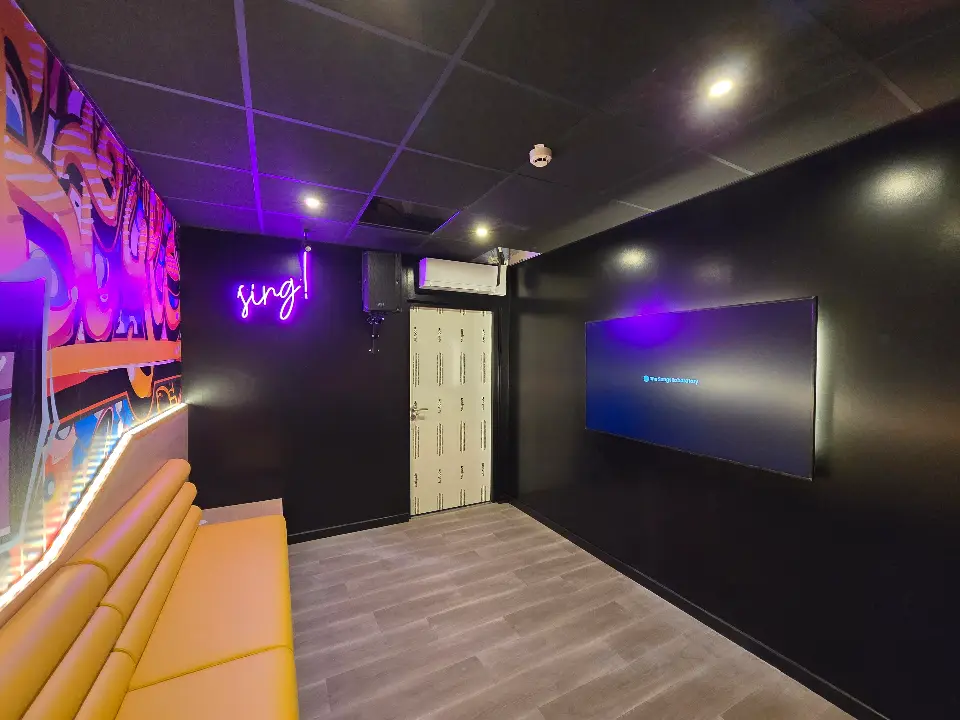 Intérieur de salle de karaoké avec un banc jaune, une fresque murale colorée, et une enseigne néon "sing!".