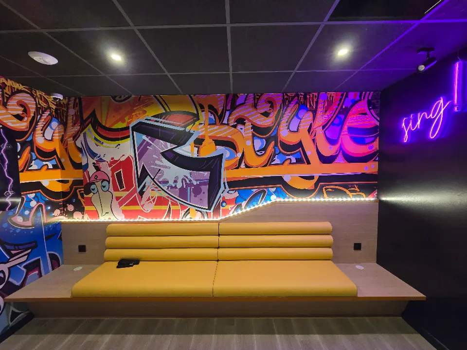 Banque jaune moutarde et longue table en bois clair face à un mur graffé, avec un néon violet "sing!".
