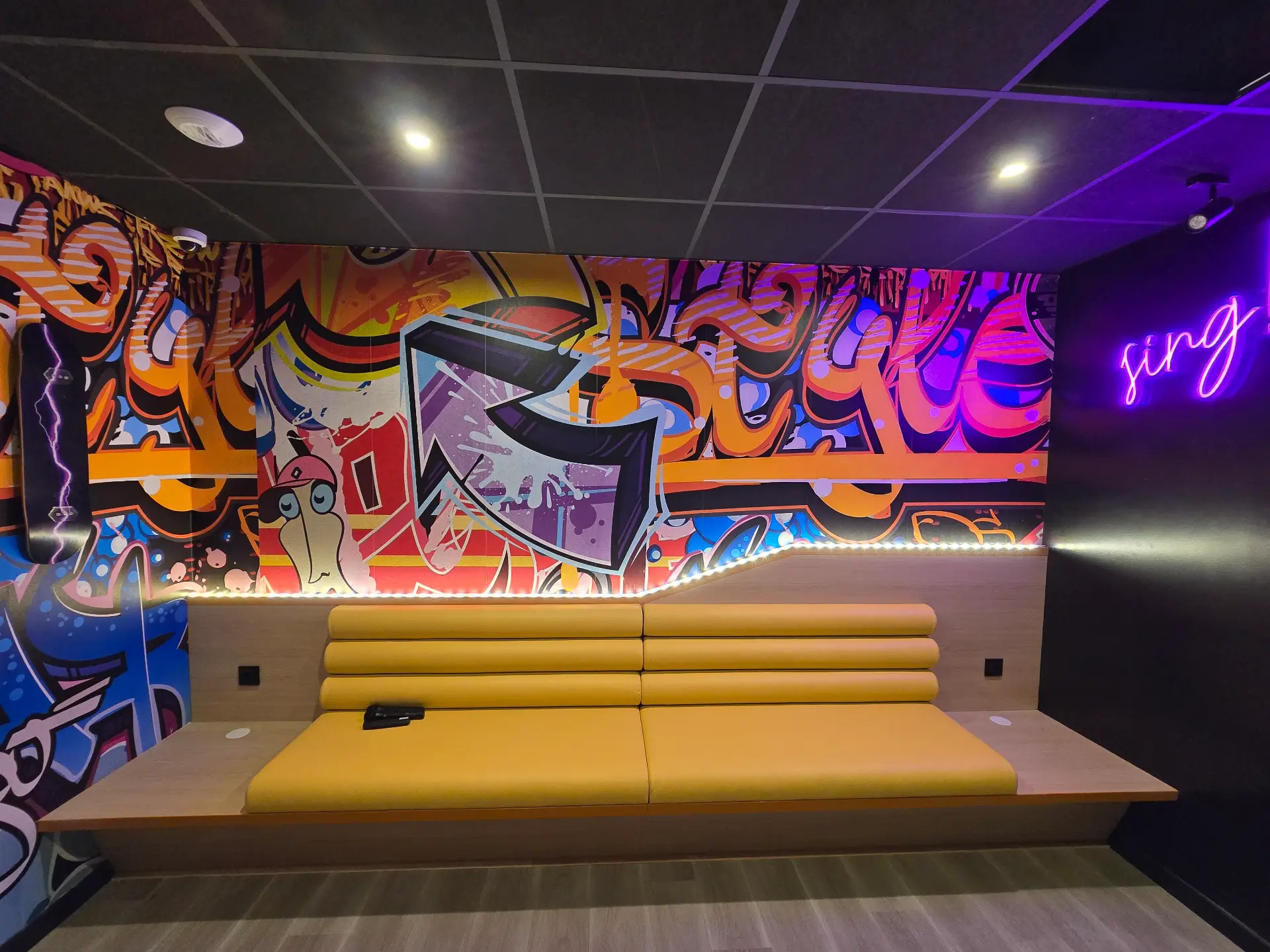 Vue en angle sur un canapé jaune moutarde encastré sur des murs colorés de style graffitis. Lumières LED sous l'étagère en bois.