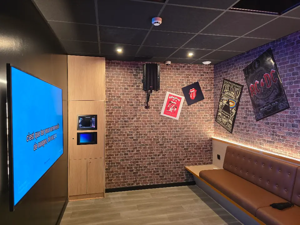 Salle de karaoké moderne avec mur en briques, écran lumineux affichant des paroles, banquette en cuir brun et affiches de groupes de rock.