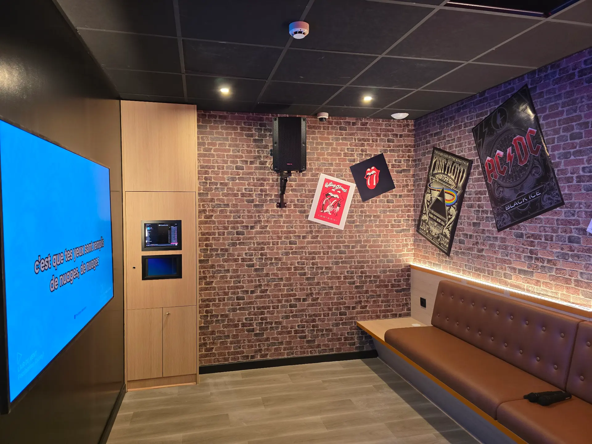 Salle de karaoké avec un canapé en cuir marron, mur en briques, posters rock, plafond sombre et enceinte de son côté droit.
