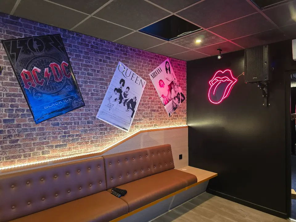 Banquette en similicuir marron dans un espace de karaoké avec des murs en briques apparentes, des affiches de groupes de rock encadrées et un néon rouge en forme de bouche, éclairage d'ambiance.