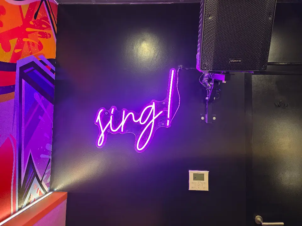 Néon "Sing!" violet vif sur mur sombre, haut-parleur noir et panneau mural coloré, lumière d'ambiance.
