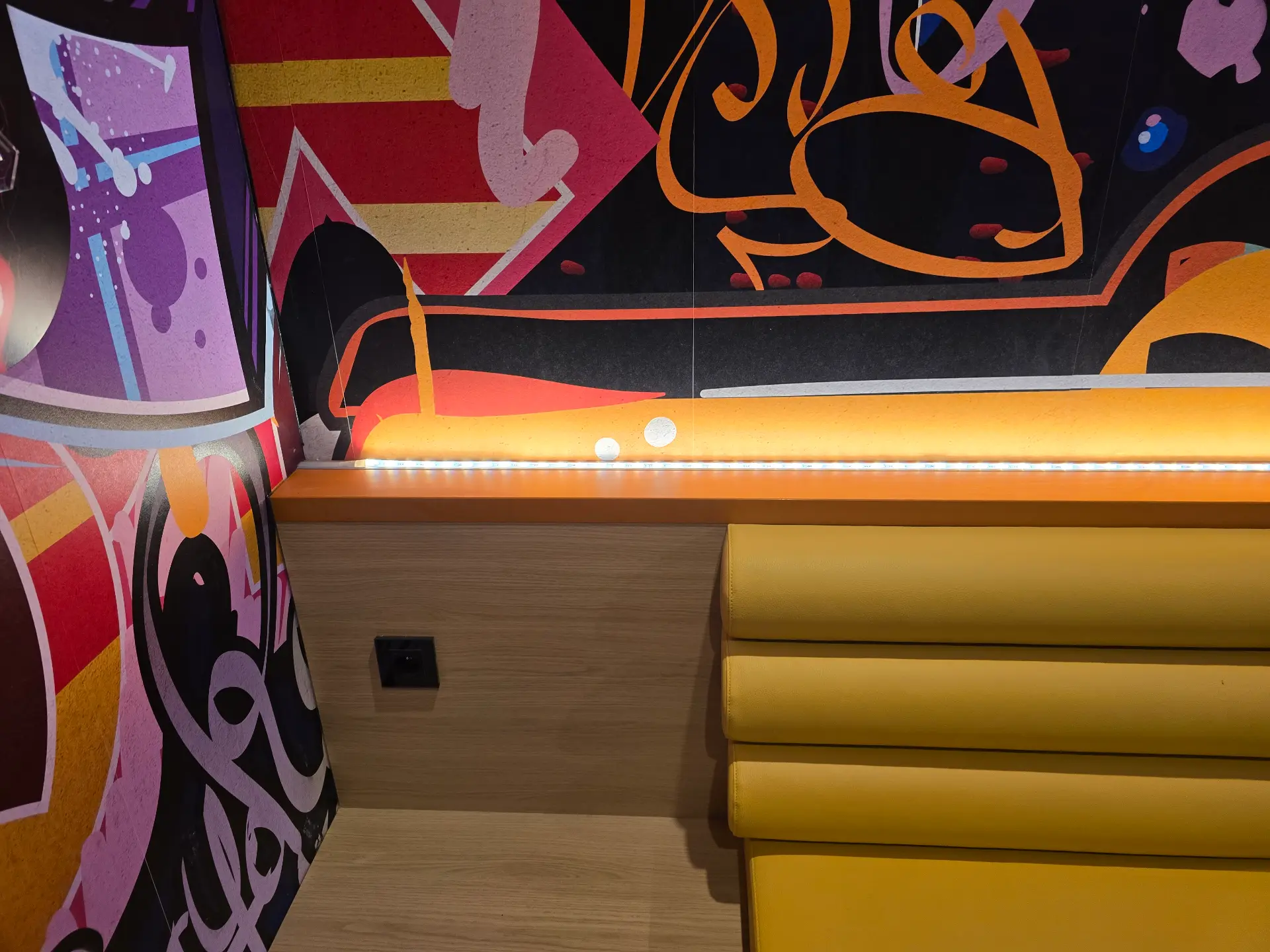 Rembourrage de banquette jaune moutarde et mur boisé clair, éclairés par une bande lumineuse LED, avec des motifs graffiti.